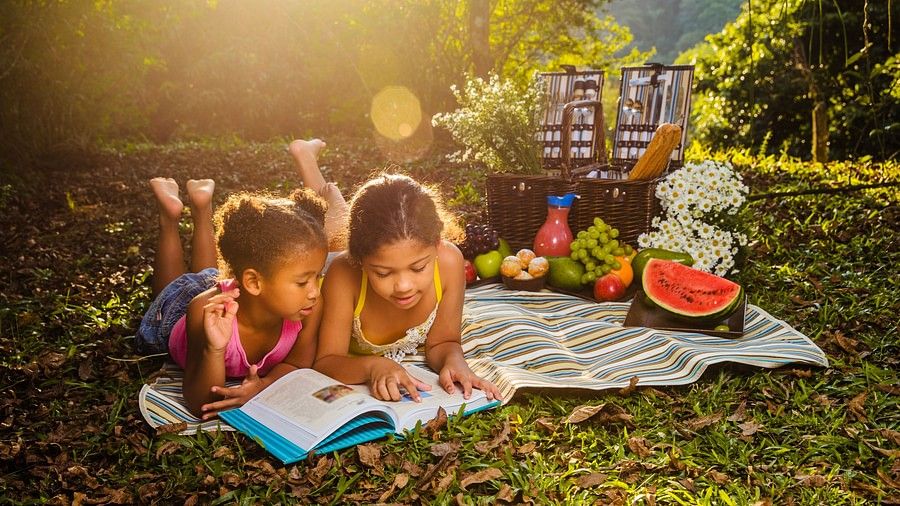 Two kids laying down to read a book on a blanket on the grass, while on a picnic